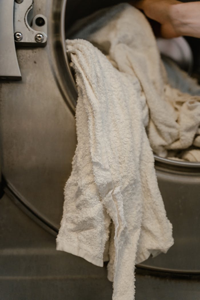 Home White towels partially hanging from a stainless steel washing machine in a laundromat.